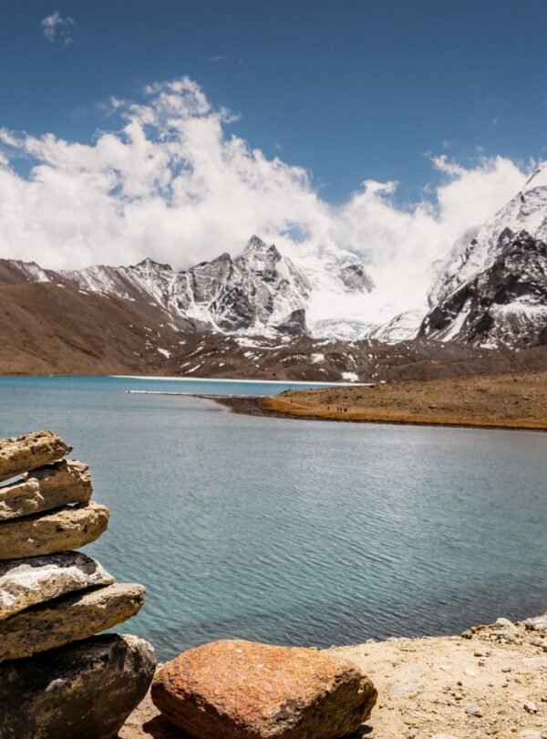 Tsongmo Lake in East Sikkim with crystal clear turquoise waters reflecting snow-capped Himalayan peaks. Stacked stone cairn in foreground overlooks the high-altitude alpine lake surrounded by brown terrain and majestic snow-dusted mountains under bright blue skies with scattered clouds. Popular tourist destination showcasing Sikkim's breathtaking natural landscape at high elevation.
