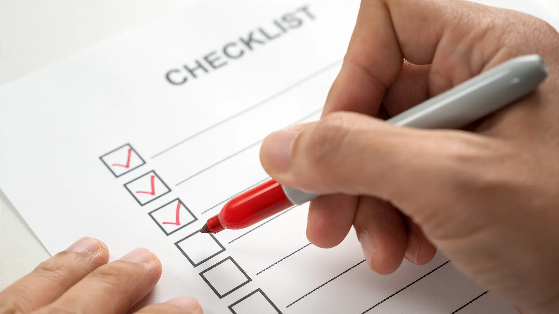 Hand holding red marker checking boxes on a printed checklist paper.