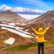 Trekker in a yellow jacket overlooks the dramatic snow-patched brown mountains and towering Himalayan peaks of Sikkim under a clear blue sky, showcasing Sikkim’s natural beauty and ideal trekking destination.